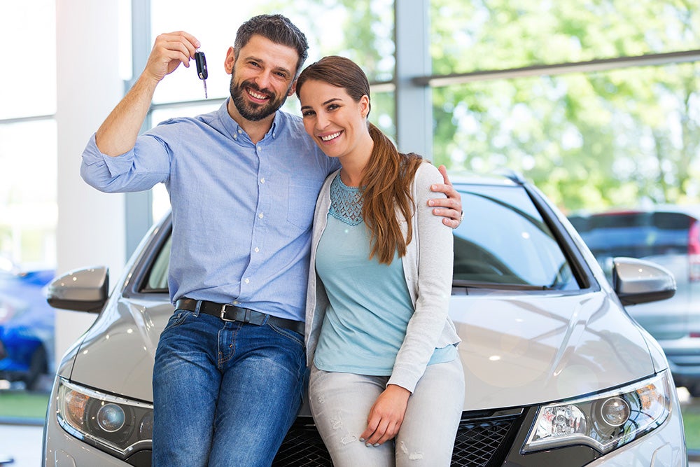 A smiling couple leans against their new car while holding up keys
